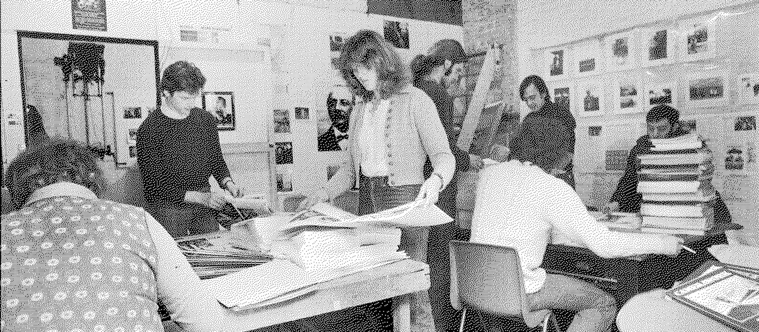 Black and white photograph depicting a person working at a drafting table. The individual is placing materials on a large sheet of paper under the illumination of an adjustable desk lamp. The workspace is cluttered with drafting tools and papers, suggesting an active, creative environment. The background features additional office supplies and decor, indicating a design or artistic studio setting.