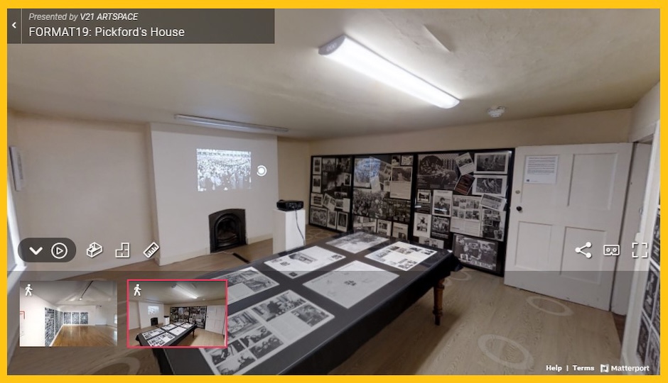 Exhibition room at Pickford's House showcasing archival photographs and documents on display. The image shows a rectangular table, with numerous historical prints and papers spread across its surface, under fluorescent lighting. To the right, a large display board on the wall features a collage of black-and-white photographs and documentation. Above the fireplace, a projection is visible on the wall. The room has a minimalist design with light-colored walls and wood flooring.