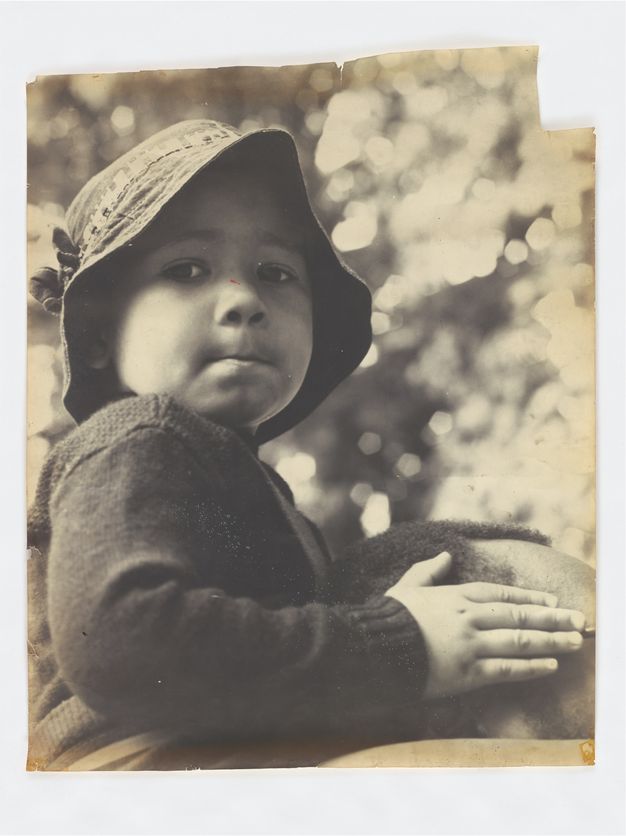 Black and white photograph depicting a young child wearing a hat, looking towards the camera. The child is wearing a knitted sweater and has one hand resting on an adult's shoulder, suggesting a sense of trust and security. The background is softly blurred, emphasizing the child's contemplative expression. The edges of the photograph are slightly worn, adding a vintage feel to the image.