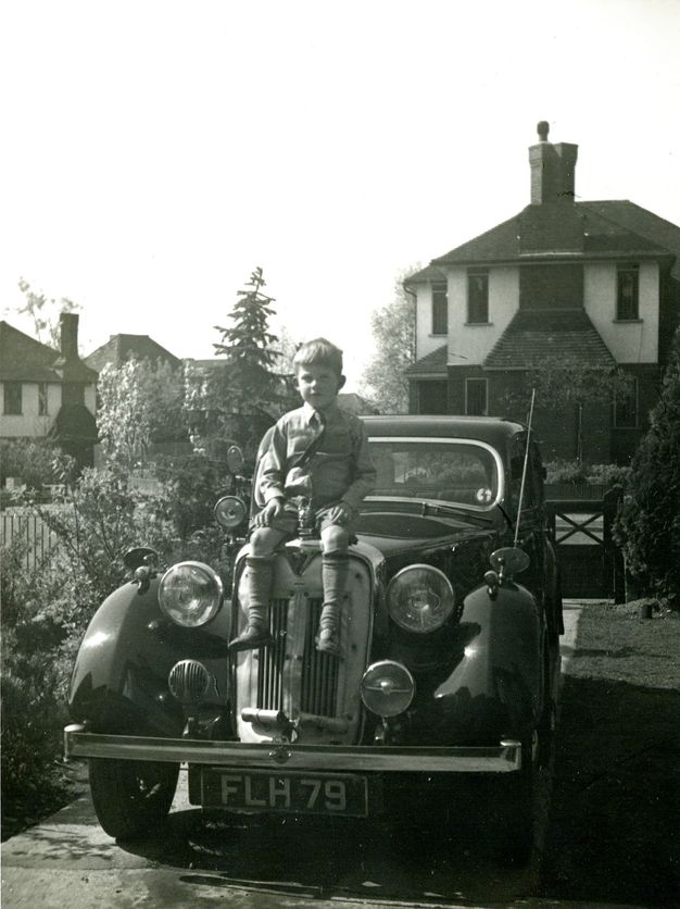 Black and white photograph of a young boy seated on the front bumper of a vintage car with the license plate "FLH 79." The car is parked in a residential driveway in front of a two-story house. The background includes other similar houses, trees, and a garden area.