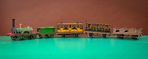 A model train set on a bright green surface against a brown background. The train set comprises five segments: a green steam locomotive, a green tender marked "G.W.J.R.," a yellow passenger carriage, a black passenger carriage, and a brown open freight car numbered "3."