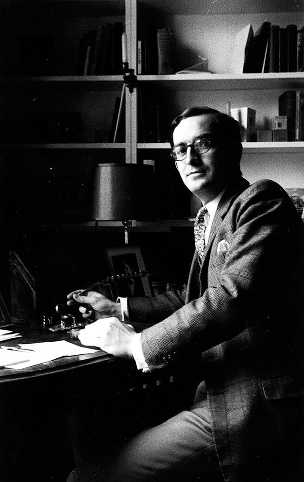 A black-and-white photograph of a man seated at a desk in an office setting. He is dressed in a suit and tie, wearing glasses, and is looking towards the camera. He holds a feather pen. On the desk are papers, a lamp, an obscured photograph, and an ink bottle.