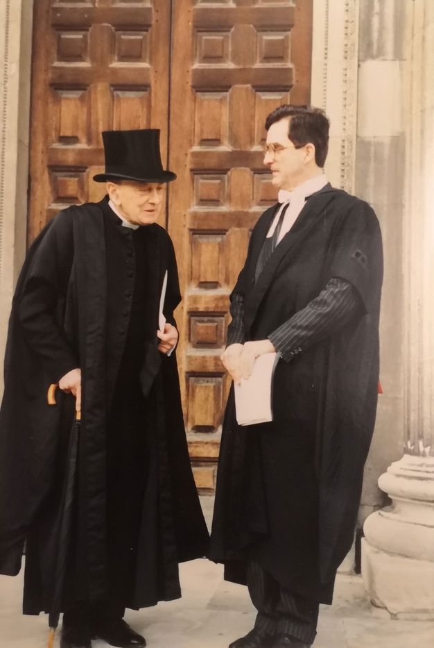 Two men in black and white formal academic attire stand conversing outside a large wooden door. The man on the left, wearing a top hat and holding a cane, appears to be elderly. The man on the right wears glasses and holds a document.