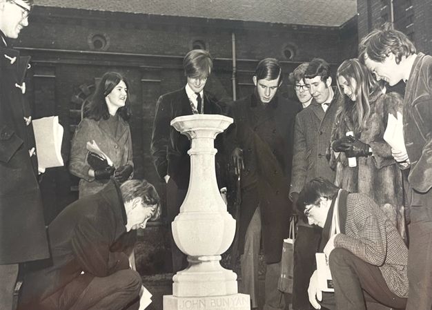 In this black and white photograph a group of young adults gather around a stone pedestal with "John Bunyan" inscribed at its base. They wear suits or wool coats and appear to examine or discuss the object of interest.