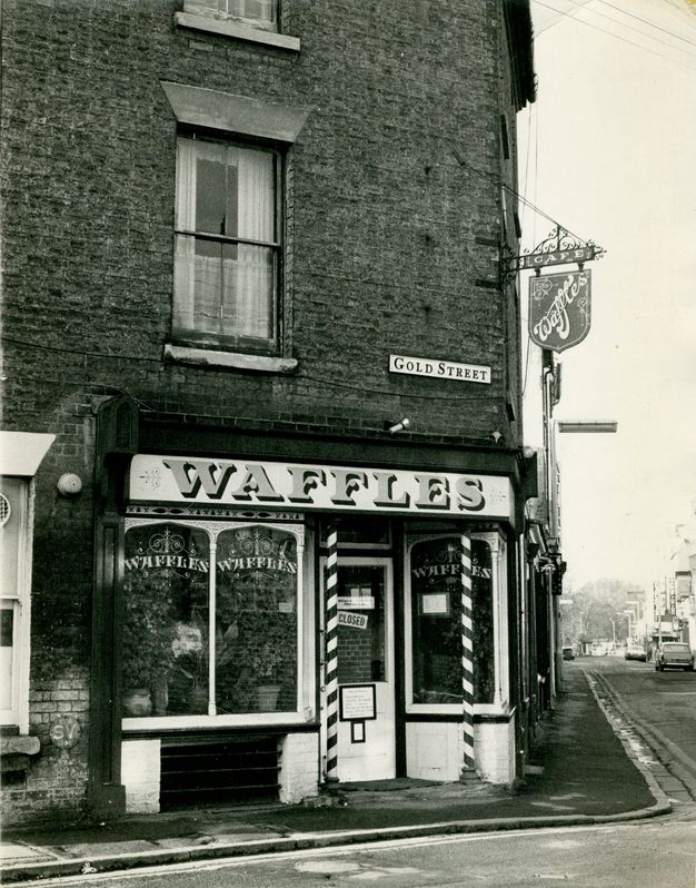 Black and white photograph of a brick café on a street corner under a small sign that reads "Gold Street". A hanging sign in the shape of a crest reads "Café Waffles". The shop front has three glass windows, each with the word "waffles" printed on it. Two narrow spiral-stripped columns frame the door.