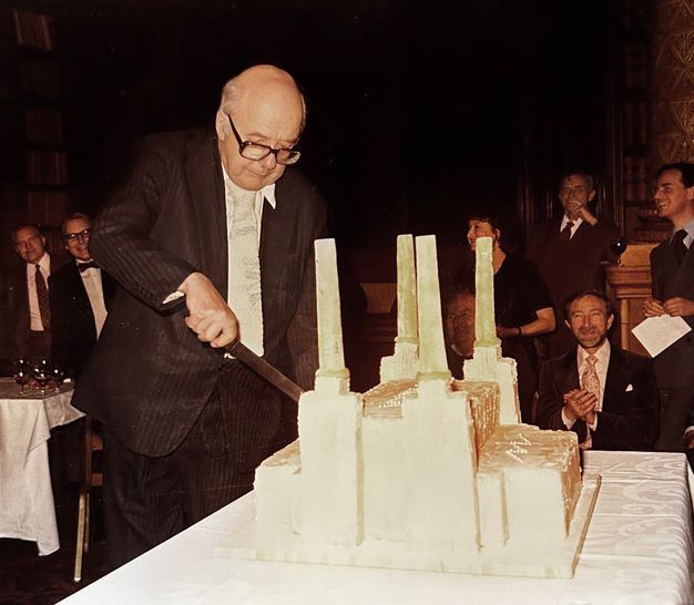 An elderly man in a suit and tie is cutting into a large, rectangular cake designed to resemble a building with four tall smokestacks. The event is set indoors with a dimly lit, formal setting. Several individuals in suits and formal attire are gathered around, some seated and clapping, suggesting a celebratory occasion. The room appears to be decorated in a classical style with ornate patterns on the walls.