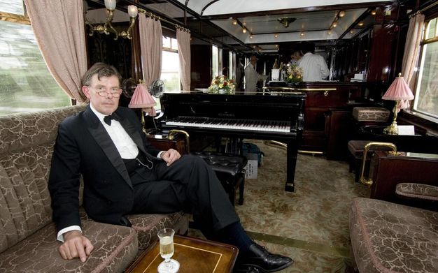 A man in formal attire, wearing a black tuxedo and bow tie, is seated on an upholstered sofa in an opulent train carriage. The setting features luxurious wooden paneling, floral arrangements, and soft lighting from decorative lamps. A black grand piano with the brand "Schimmel" is prominently displayed in the background. The man holds a composed expression and a glass of champagne rests on a small table in front of him. Plush curtains line the windows, enhancing the elegant atmosphere of the interior.