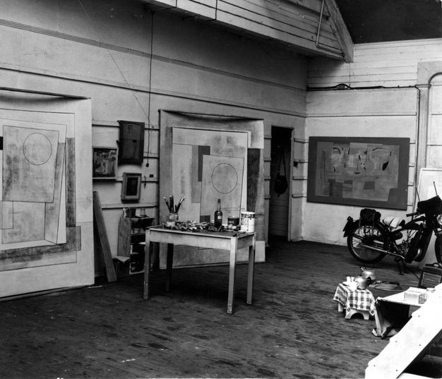 Black and white photograph of an artist's studio featuring large abstract paintings with geometric shapes on the walls. A small rectangular table is covered with brushes and bottles. There is a motorcycle against the back wall.