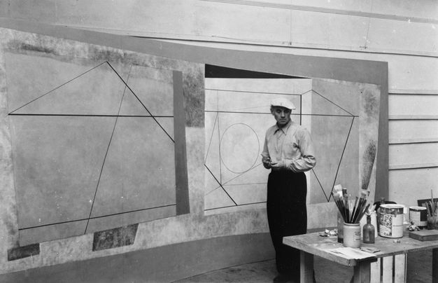 Black and white photograph of an artist standing in a studio, examining large-scale geometric abstract paintings mounted on the wall. The paintings feature intersecting lines and shapes, including triangles and circles. The artist, wearing a light-colored shirt and cap, is positioned beside a table filled with paintbrushes and art supplies. The setting appears to be an art studio, with the focus on the creative process and environment of abstract art practice.