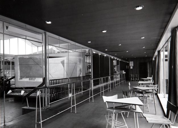 Black and white photograph of an interior corridor in a modernist building, featuring a geometric mural on a central partition wall with abstract shapes and lines. The space is accented by a series of pendant light fixtures on the ceiling and is bordered by glass panels on one side, allowing natural light to enter. A series of minimalist wooden chairs and tables are arranged along the corridor, with curtains lining the glass wall. The floor is polished, reflecting the streamlined aesthetic of mid-20th century architecture.