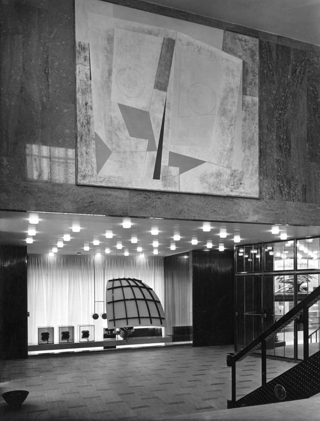 Black and white photograph of a modern interior space, possibly part of a building lobby. The composition features a large abstract mural on the dark marble wall above, characterized by geometric shapes and subtle textural contrasts. Beneath the mural, a display area is illuminated by a grid of recessed ceiling lights. The display includes a large, semi-circular dome structure, resembling a segmented globe, complemented by minimalist decor and three framed images or artworks on the left.